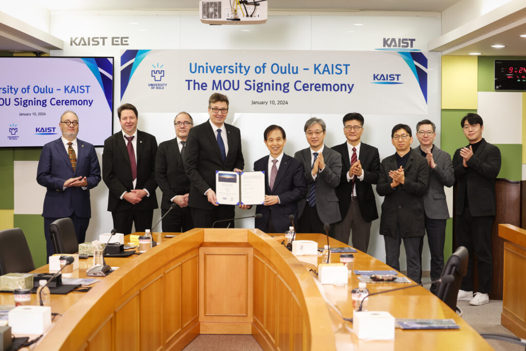 A group of dignitaries from the University of Oulu and KAIST stands behind a conference table, applauding as two central figures hold up a signed memorandum of understanding.