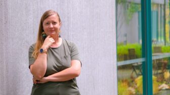 Doctoral researcher Anna Teern stands against a pale concrete wall with her hand to her chin, beside a green-framed window.