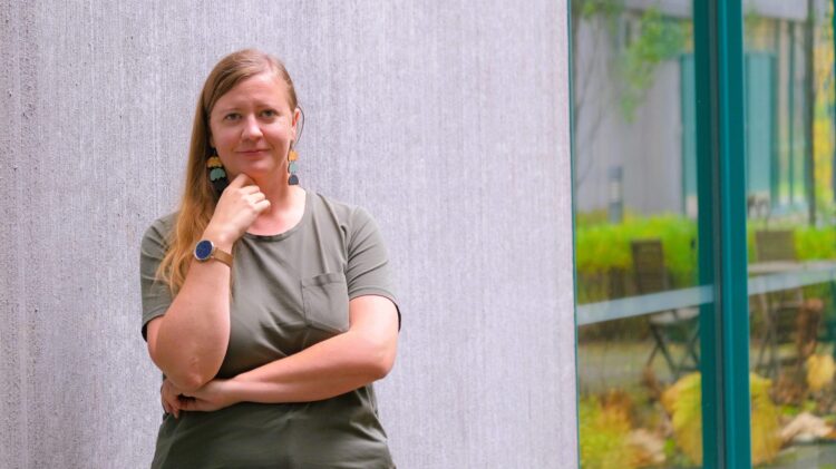 Doctoral researcher Anna Teern stands against a pale concrete wall with her hand to her chin, beside a green-framed window.