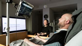 Harri Pennanen, a researcher with ALS, lies in a hospital bed in his apartment using an eye-tracking computer to work, with Tuomo Hänninen sitting beside him. A screen before Harri displays the title of his published 6G paper in IEEE Access.