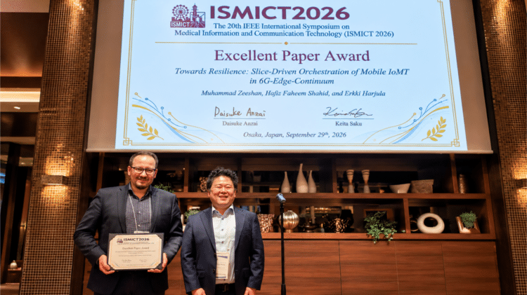 Erkki Harjula (on the left) holding the Excellent Paper Award certificate at ISMICT 2026 in Osaka, with the award slide on screen behind him.