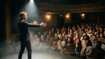 A performer on a theatre stage extends his hand toward a captivated audience, with spotlight beam and low smoke filling the foreground.