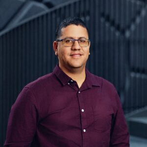 Osmel Martínez in glasses wearing a maroon shirt standing in front of a dark architectural background