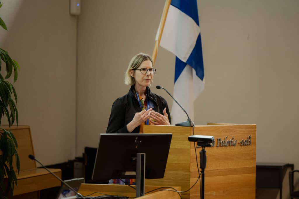 Sabine Koch speaking at a lectern beside the Finnish flag